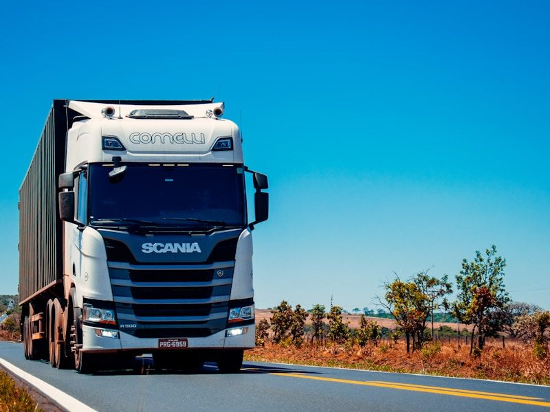 White prime mover hauling a container trailer along a two-lane highway through dry scrub